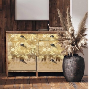 May include: A wooden dresser with six drawers, each with a gold and white floral pattern. The dresser has black metal handles. The dresser is sitting on a wooden floor. A vase with dried pampas grass is in the foreground.