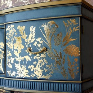 May include: A dark blue cabinet featuring a gold botanical pattern of various wildflowers and grasses across the front panel. The piece includes a curved gold metal handle centred on the drawer and gold trim along the edges and base.