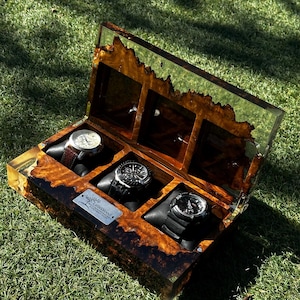 Puede incluir: Una caja de madera para relojes con tres compartimentos, que muestra tres relojes diferentes. La caja tiene una tapa transparente, similar a la resina, y un rico patrón de vetas de madera marrón. La caja está abierta, revelando los relojes sobre cojines negros, sobre hierba verde.