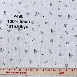 May include: White linen fabric with a delicate floral pattern in shades of blue and green. The fabric has the text "#490" and "100% linen" printed on it. A wooden ruler is at the bottom of the image.