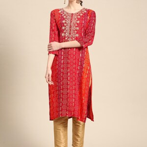 Puede incluir: Una kurta de estampado rojo y naranja con bordados dorados. La kurta tiene un escote redondo y mangas tres cuartos. La modelo lleva pantalones dorados y zapatos marrones.