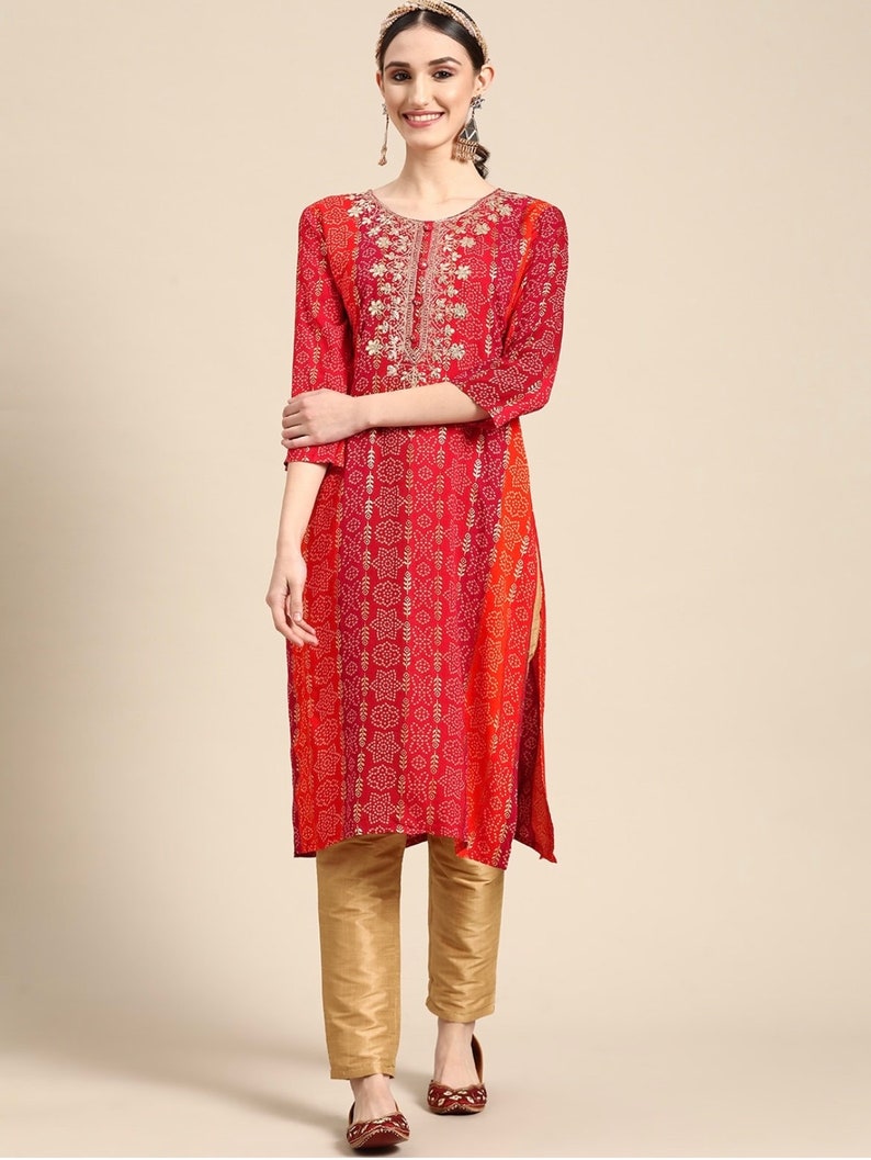 Puede incluir: Una kurta de estampado rojo y naranja con bordados dorados. La kurta tiene un escote redondo y mangas tres cuartos. La modelo lleva pantalones dorados y zapatos marrones.