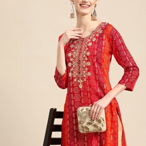 Puede incluir: Una kurta de manga larga roja y naranja con bordados dorados y un clutch blanco. La kurta tiene un patr&oacute;n impreso de peque&ntilde;os puntos blancos y un dise&ntilde;o floral m&aacute;s grande.