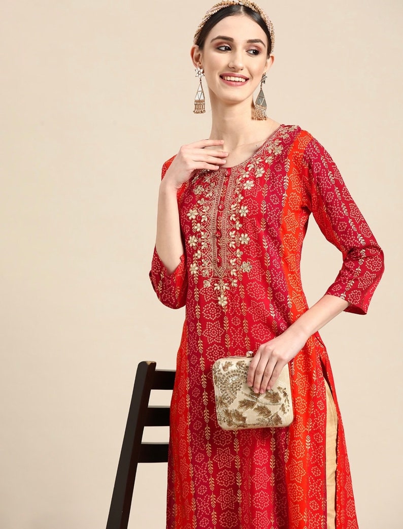 Puede incluir: Una kurta de manga larga roja y naranja con bordados dorados y un clutch blanco. La kurta tiene un patr&oacute;n impreso de peque&ntilde;os puntos blancos y un dise&ntilde;o floral m&aacute;s grande.