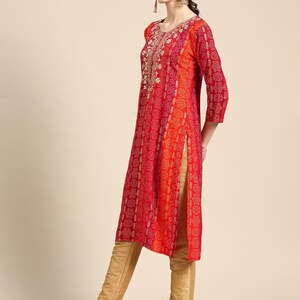 Puede incluir: Una kurta de manga larga roja y naranja con bordados dorados y estampado floral. La kurta tiene un escote redondo y una abertura lateral. La modelo lleva la kurta con pantalones dorados y zapatos rojos.