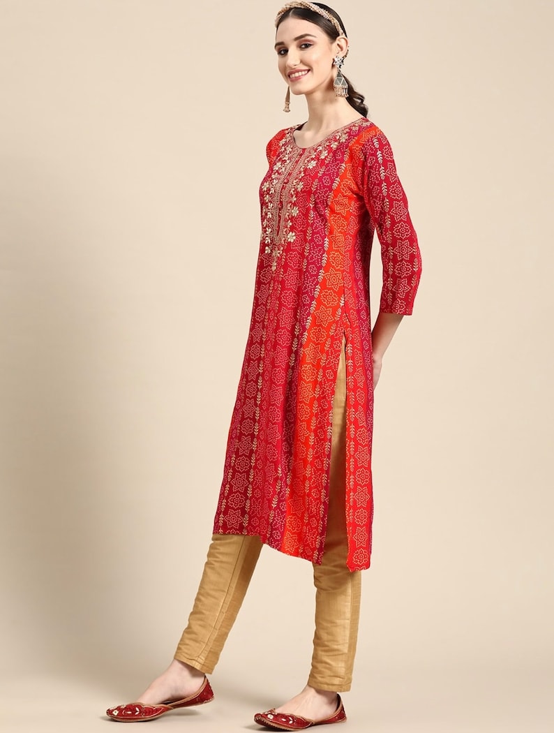 Puede incluir: Una kurta de manga larga roja y naranja con bordados dorados y estampado floral. La kurta tiene un escote redondo y una abertura lateral. La modelo lleva la kurta con pantalones dorados y zapatos rojos.