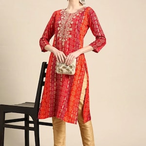 Puede incluir: Una kurta de manga larga roja y naranja con un cuello bordado en oro y una abertura lateral. La kurta se combina con pantalones dorados y zapatos rojos.