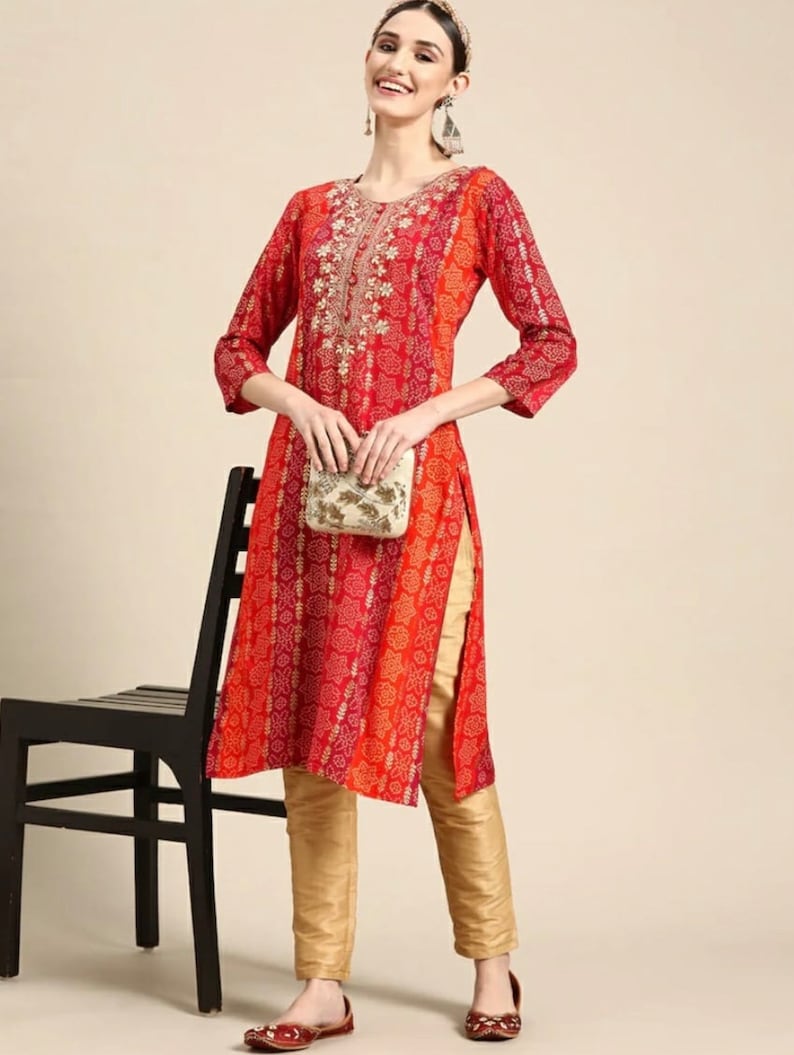 Puede incluir: Una kurta de manga larga roja y naranja con un cuello bordado en oro y una abertura lateral. La kurta se combina con pantalones dorados y zapatos rojos.