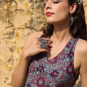 May include: A woman wearing a sleeveless dress with a floral print in shades of pink, blue, and green. She is wearing a silver ring with a butterfly design and silver earrings.
