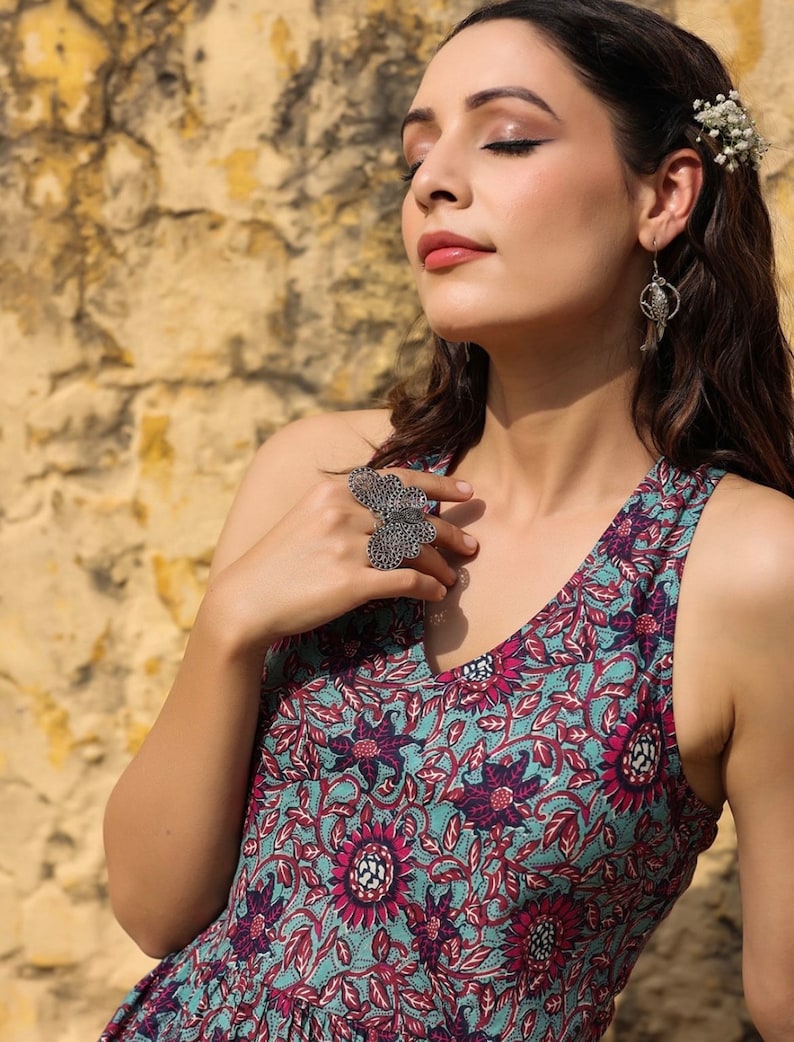 May include: A woman wearing a sleeveless dress with a floral print in shades of pink, blue, and green. She is wearing a silver ring with a butterfly design and silver earrings.