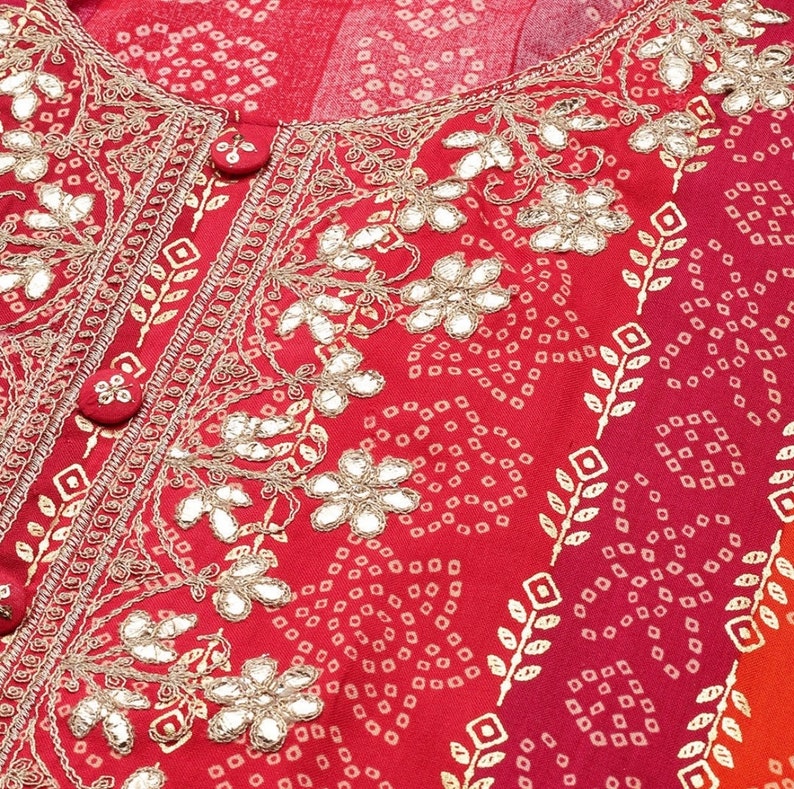 Puede incluir: Tela roja con un dise&ntilde;o bordado en oro con motivos florales y peque&ntilde;os adornos de espejo. La tela tiene un patr&oacute;n repetitivo de peque&ntilde;os puntos blancos.
