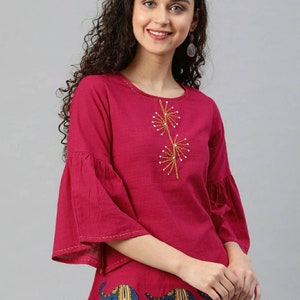 Könnte beinhalten: Ein rosa Baumwoll-Oberteil mit Glockenärmeln und gestickten Blumendetails. Der untere Teil des Oberteils zeigt einen blauen und gelben Elefanten-Druck.