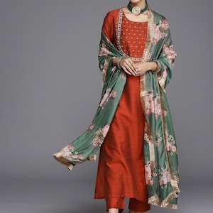 May include: A rust-colored kurta with a floral print dupatta. The kurta has a round neckline and is embroidered with gold thread. The dupatta is a light green color with pink and white floral designs.