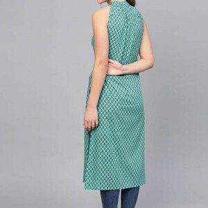 May include: A teal blue sleeveless dress with a high neck and a geometric pattern. The dress is knee-length and has a side slit. The model is wearing the dress with blue jeans and flat shoes.