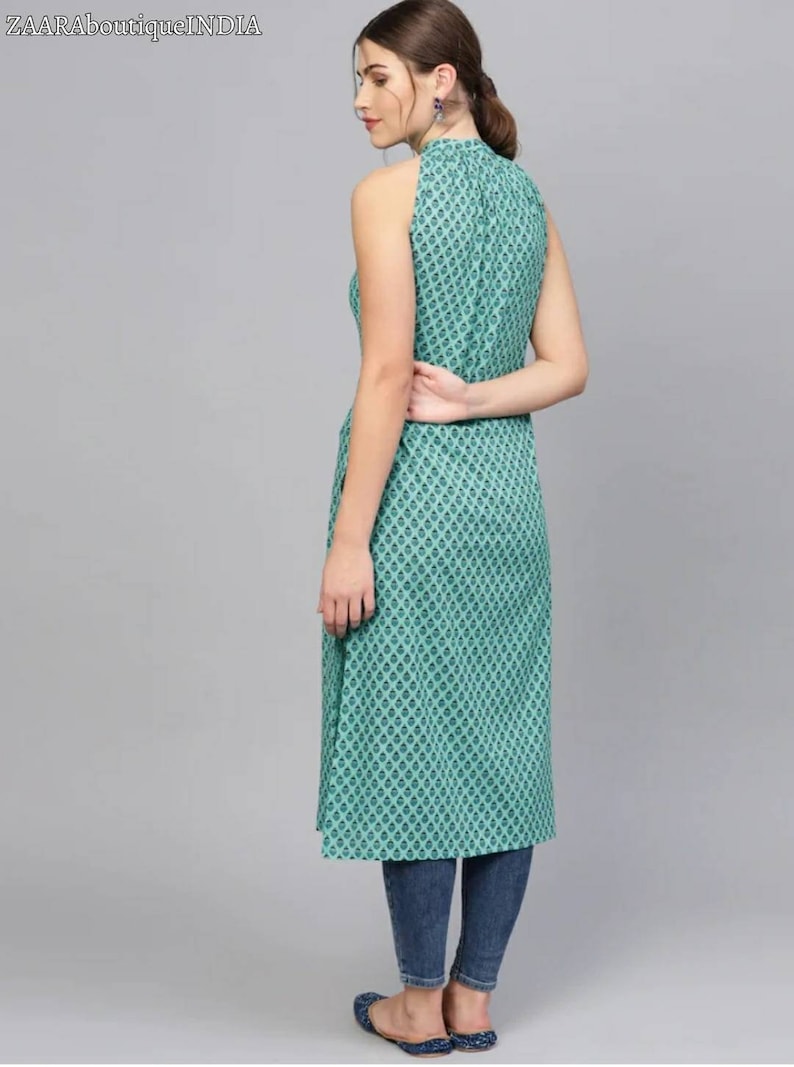 May include: A teal blue sleeveless dress with a high neck and a geometric pattern. The dress is knee-length and has a side slit. The model is wearing the dress with blue jeans and flat shoes.