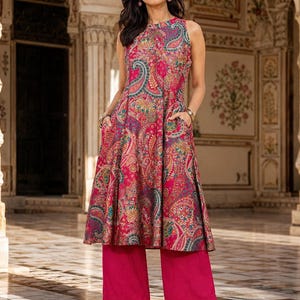May include: A sleeveless knee-length tunic featuring a vibrant pink base with a multi-colored paisley print, a round neckline, and side pockets, paired with solid pink wide-leg trousers and light pink strappy block-heeled sandals.