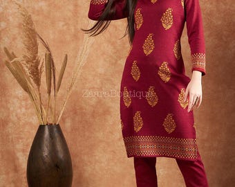 May include: A burgundy tunic and trouser set with gold floral patterns. The tunic has a mandarin collar and long sleeves with decorative gold borders on the cuffs. The hemline also features a gold border. The outfit is made of a soft, textured fabric.