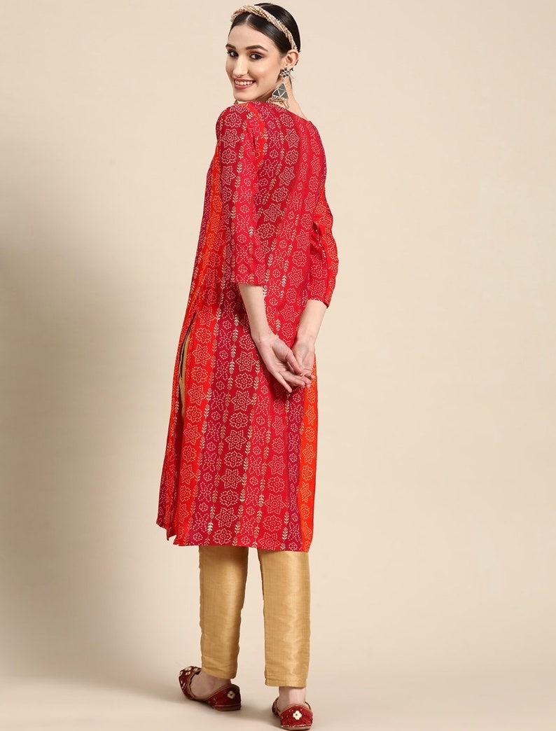 Puede incluir: Una blusa de manga larga roja y naranja con estampado floral y detalles dorados. La blusa tiene una abertura lateral y se combina con pantalones dorados.