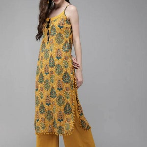 May include: A mustard yellow sleeveless kurta with a floral print and black tassel detail. The kurta has a side slit and a pom-pom trim. The model is wearing the kurta with mustard yellow palazzo pants.