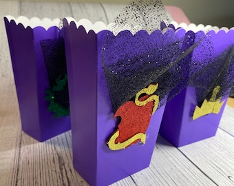 Pode incluir: Três caixas de pipoca roxas com bordas festonadas. Cada caixa tem um personagem diferente do filme da Disney Descendants. A caixa da esquerda tem um personagem verde, a caixa do meio tem um personagem vermelho com detalhes dourados, e a caixa da direita tem um personagem dourado.