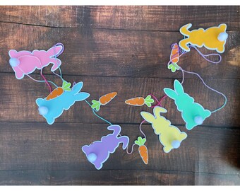 May include: A colorful Easter bunny garland with six bunnies in pastel colors: pink, blue, yellow, green, and purple. Each bunny has a white pom-pom tail and is connected by string to orange carrot cutouts. The garland is set against a dark wood background.