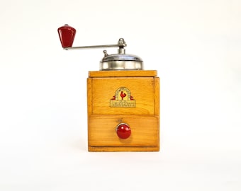May include: A vintage wooden coffee grinder with a red handle and a metal grinder. The grinder has a drawer and a label that says "Lehnartz".