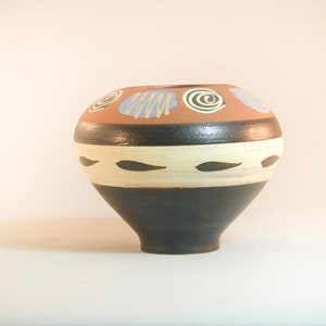 May include: A round ceramic vase with a black, white and brown geometric design. The vase has a wide base and a narrow neck.