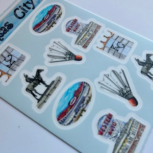 Pode incluir: Uma folha de adesivos com v&aacute;rios marcos e s&iacute;mbolos de Kansas City, Missouri. Os adesivos s&atilde;o impressos em papel branco com fundo azul claro. Os adesivos incluem um est&aacute;dio, um volante, uma est&aacute;tua de cavalo e um edif&iacute;cio com o texto "Western Auto" nele.