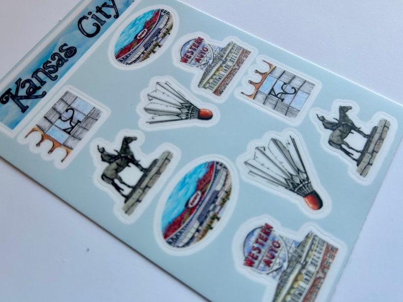 Pode incluir: Uma folha de adesivos com v&aacute;rios marcos e s&iacute;mbolos de Kansas City, Missouri. Os adesivos s&atilde;o impressos em papel branco com fundo azul claro. Os adesivos incluem um est&aacute;dio, um volante, uma est&aacute;tua de cavalo e um edif&iacute;cio com o texto "Western Auto" nele.