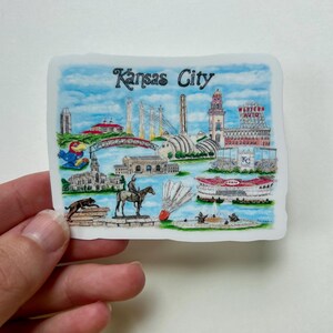 Puede incluir: Una ilustraci&oacute;n colorida de monumentos de Kansas City, incluyendo el Country Club Plaza, el Museo de Arte Nelson-Atkins, el Centro Kauffman para las Artes Esc&eacute;nicas y el Sprint Center. La ilustraci&oacute;n tambi&eacute;n incluye una representaci&oacute;n del logotipo de los Kansas City Chiefs, un volante y un jinete a caballo.