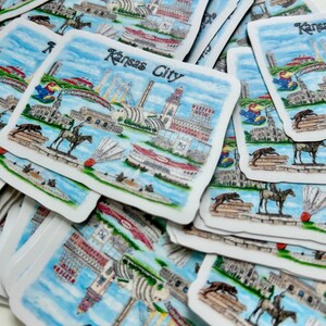Puede incluir: Una pila de pegatinas de recuerdo de Kansas City de colores que representan monumentos y atracciones. Las pegatinas est&aacute;n impresas en papel blanco con un acabado brillante.