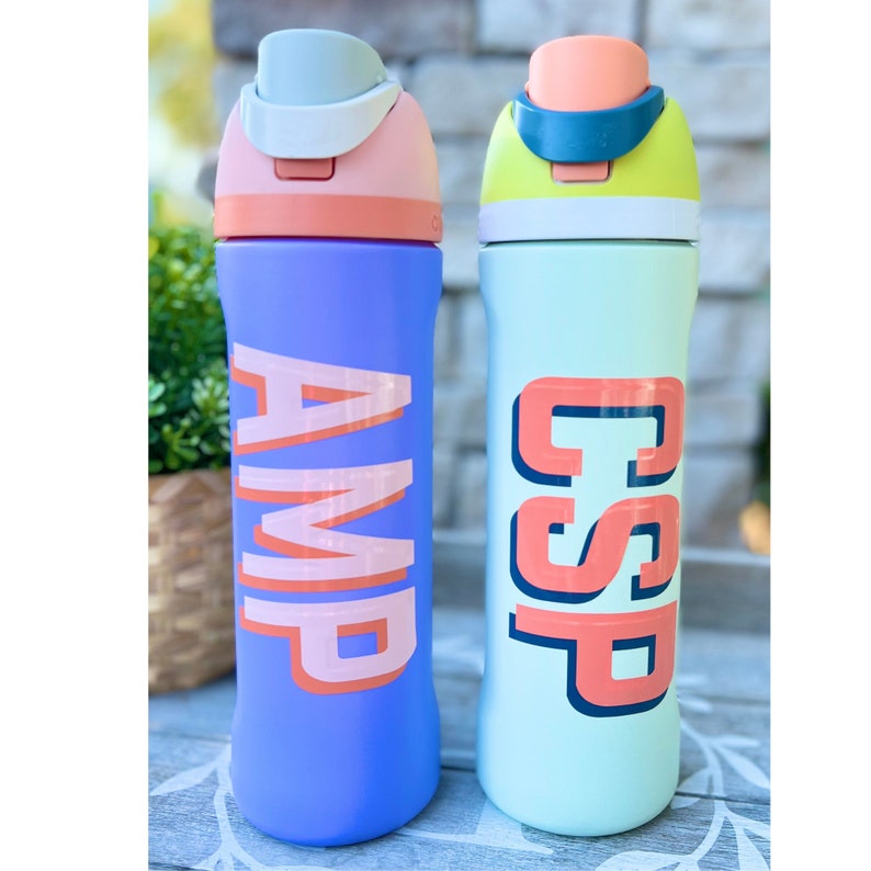 Może przedstawiać: Dwie kolorowe butelki z wodą z personalizowanymi nazwami "AMP" i "CSP" w retro stylu czcionki. Butelki są niebieskie i zielone z r&oacute;żowymi i ż&oacute;łtymi akcentami.