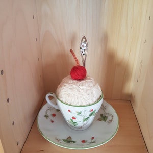 May include: A white crocheted pincushion shaped like a cupcake with a red yarn cherry on top, sitting in a white teacup with a green and white floral pattern on a white saucer with a green rim.