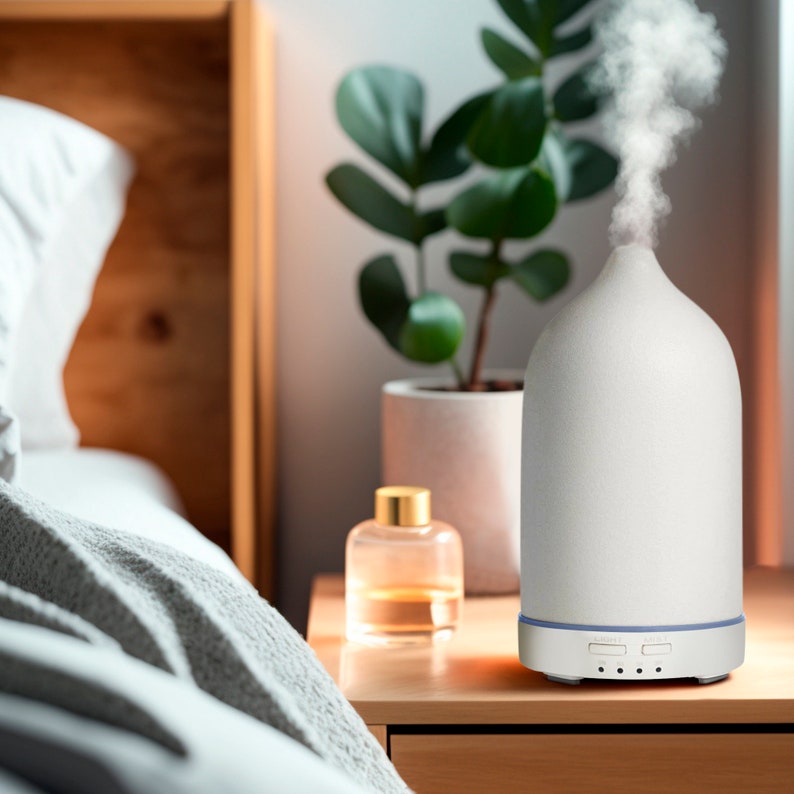 May include: White ceramic essential oil diffuser with a round base and a dome-shaped top. The diffuser is on a wooden table next to a clear glass bottle and a green plant.