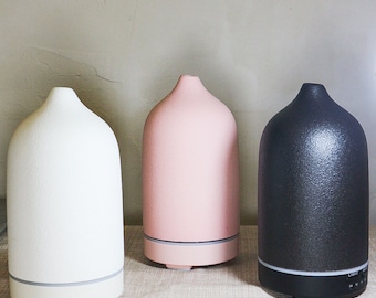 May include: Three ceramic essential oil diffusers in white, pink, and black. The diffusers have a rounded, bell-shaped design with a textured surface.
