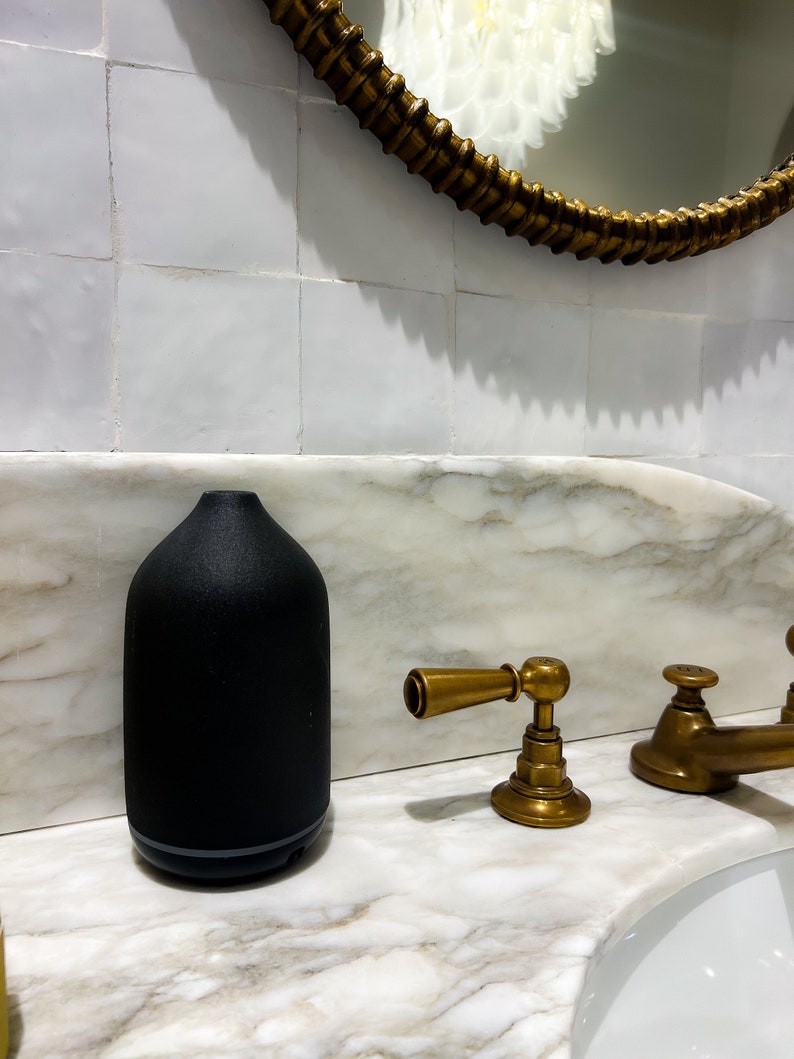 May include: A black matte ceramic essential oil diffuser sits on a white marble countertop next to a gold faucet.
