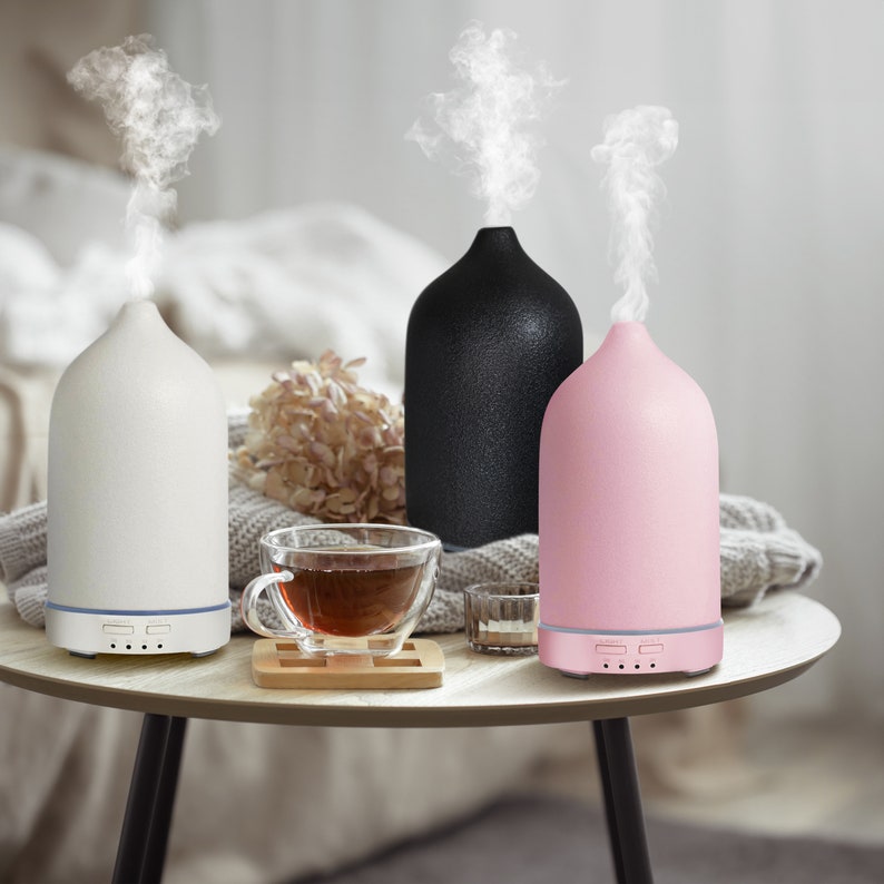 May include: Three ceramic essential oil diffusers in white, black, and pink. The diffusers are on a wooden table with a cup of tea.