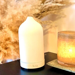 May include: White ceramic essential oil diffuser with the word "Ara" printed on the bottom. The diffuser is sitting on a dark wooden surface next to a glass candle holder.