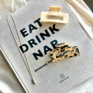 May include: A book with the text "Eat Drink Nap" and the words "Bringing the" at the bottom. Two hair clips are on the book, one is white and the other is brown with black spots. A gold chain necklace with a small pendant is on the book.
