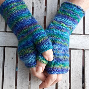 Könnte beinhalten: Gestrickte Fingerhandschuhe in Blau-, Grün- und Lilatönen. Die Handschuhe haben ein strukturiertes Muster und sind handgefertigt. Die Bündchen und Fingeröffnungen sind gerippt. Die Handschuhe sind auf einer weißen Holzoberfläche zu sehen. Die Handschuhe sind ca. 20 cm lang.