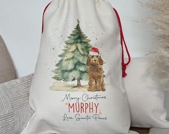 Op de afbeelding: Een gepersonaliseerde kerstcadeauzak met een rood trekkoord. De tas heeft een aquarel-illustratie van een hond met een kerstmanmuts die naast een kerstboom staat. De tekst op de tas is "Merry Christmas *Murphy* Love Santa Paws".