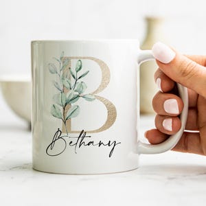 Pode incluir: Caneca de cerâmica branca com uma letra dourada "B" e um desenho de folhas verdes. O nome "Bethany" está escrito em itálico preto abaixo da letra.