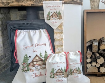 May include: A set of Christmas-themed gift bags and a banner. The bags and banner are white with red drawstrings and accents. They feature a cabin scene with the text "Special Delivery for Charlie" and "Santa please stop here for Charlie".