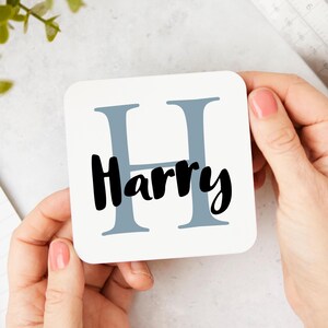 Op de afbeelding: Een witte vierkante onderzetter met een grote grijze letter H en de naam Harry eronder in het zwart.