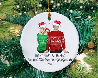 May include: A white round Christmas ornament with a drawing of a couple in red and green Christmas sweaters holding a baby. The text reads "Nanny, Henry & Grandad Our First Christmas as Grandparents 2023".