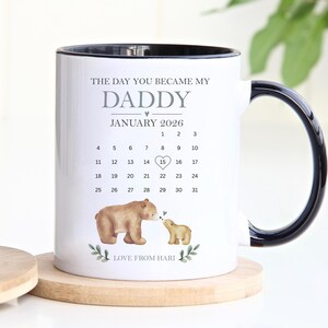 Peut inclure: Mug en céramique blanche avec une anse et un bord noirs. Le mug présente le texte "THE DAY YOU BECAME MY DADDY" et un calendrier de janvier 2026, avec le 15 mis en évidence. Comprend des illustrations d'ours et le texte "LOVE FROM HARI".