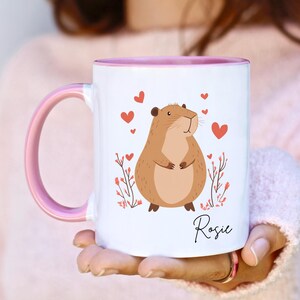 Puede incluir: Una taza de cer&aacute;mica blanca con un borde y un asa de color rosa. La taza presenta una ilustraci&oacute;n de dibujos animados de un h&aacute;mster marr&oacute;n con corazones rosas y flores. El nombre "Rosie" est&aacute; impreso en la taza.