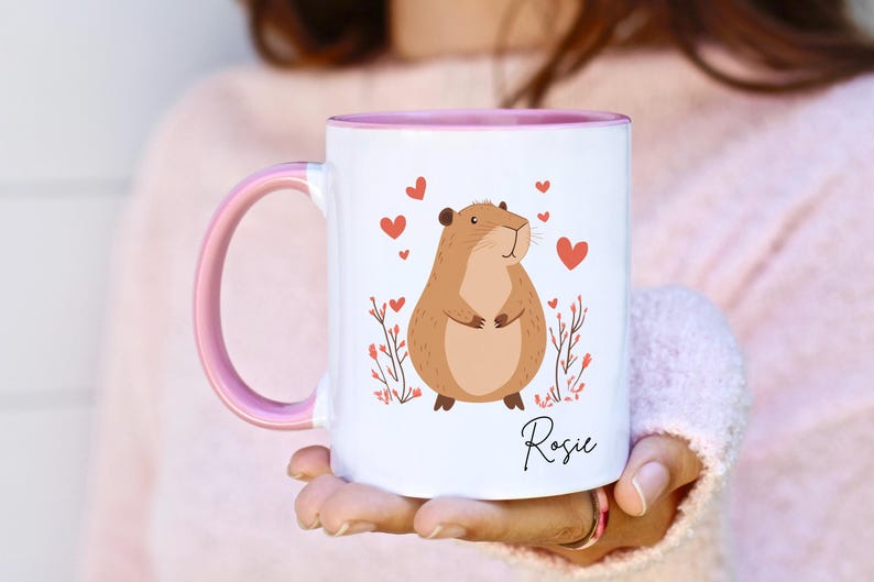 Puede incluir: Una taza de cer&aacute;mica blanca con un borde y un asa de color rosa. La taza presenta una ilustraci&oacute;n de dibujos animados de un h&aacute;mster marr&oacute;n con corazones rosas y flores. El nombre "Rosie" est&aacute; impreso en la taza.