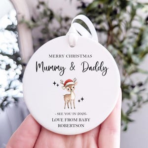 Op de afbeelding: Een witte, ronde kerstversiering met een lint. Het toont de tekst "MERRY CHRISTMAS Mummy & Daddy" en een cartoon hert met een kerstmuts. De versiering bevat ook de tekst "SEE YOU IN 2026 - LOVE FROM BABY ROBERTSON."