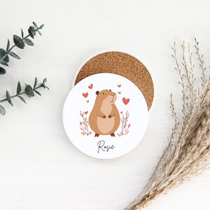 Puede incluir: Posavasos de cer&aacute;mica blanco con un h&aacute;mster de dibujos animados marr&oacute;n y blanco con corazones rojos y el nombre Rosie impreso en la parte inferior.